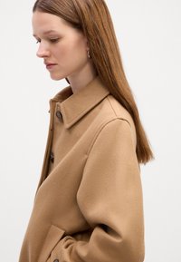 Brun uldblandingsfrakke med krave, to knapper og en struktureret finish. Model vender mod højre med langt, glat hår og øreringe.