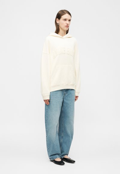 Jeune femme portant un sweat à capuche oversize de couleur blanc cassé, un jean large bleu clair et des chaussures plates noires, debout devant un fond uni.
