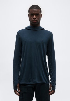 Άνδρας που φοράει σκούρο μπλε μακρυμάνικο πουκάμισο με κουκούλα και μαύρο παντελόνι, στέκεται μπροστά σε απλό φωτεινό φόντο.