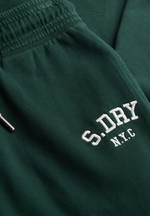 Pantalons de survêtement vert foncé avec taille élastique et logo brodé blanc "S.DRY N.Y.C" sur la cuisse près d'une petite étiquette de marque en métal.