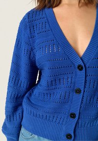 Blå stickad cardigan med djup V-ringning, strukturerad mönstring och svarta knappar. Tyget har en andningsbar, öppen vävdesign.