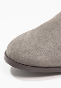 Bottine grise en daim avec un bout arrondi, une semelle plate marron et une texture lisse, mettant en avant un design minimaliste.