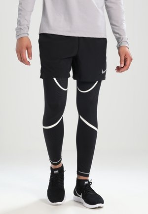 Czarne legginsy sportowe z białymi falistymi akcentami, w połączeniu z czarnymi szortami i czarnymi butami sportowymi z logo Nike. Szara koszulka z długim rękawem.