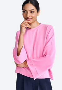 Rózsaszín, oversized pulóver, széles ujjakkal, kerek nyakkivágással és puhafa szerkezettel; sötétkék nadrággal párosítva. Kényelmes, laza szabás.