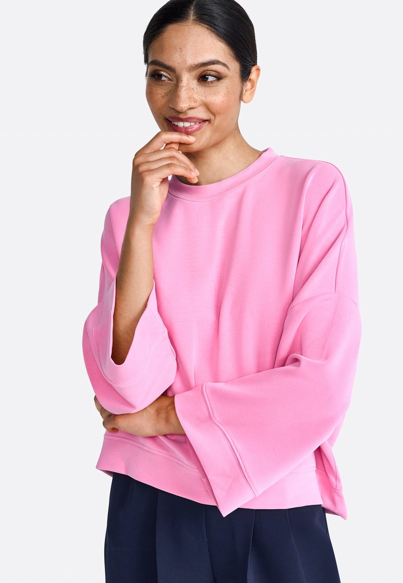 Rózsaszín, oversized pulóver, széles ujjakkal, kerek nyakkivágással és puhafa szerkezettel; sötétkék nadrággal párosítva. Kényelmes, laza szabás.