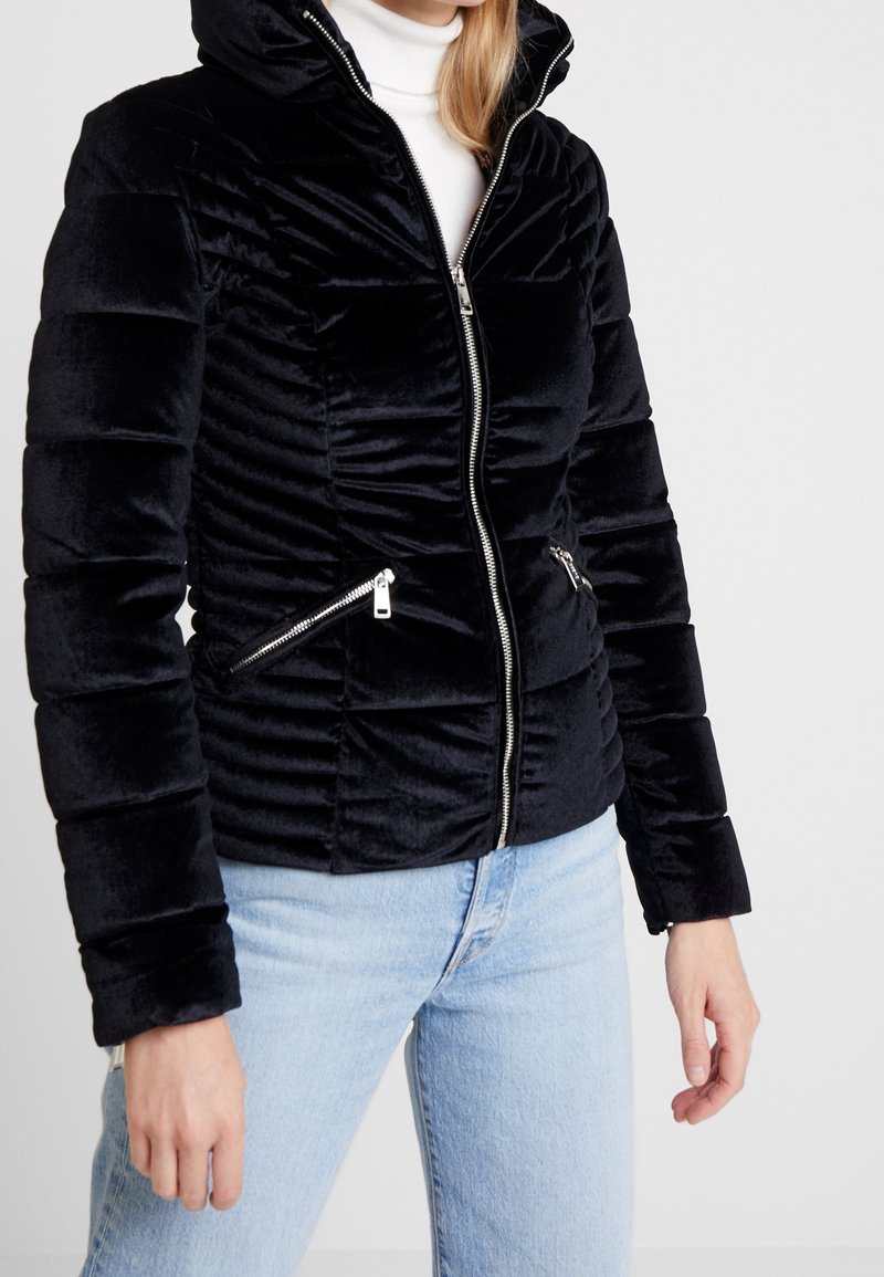 Veste rembourrée en velours noir avec fermeture éclair à l'avant, design froncé, deux poches latérales zippées et une capuche. Portée avec un jean bleu clair.