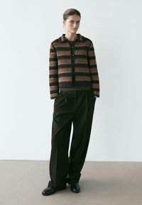 Frau steht vor einfachem Hintergrund, trägt eine braun-schwarz gestreifte, geknöpfte Strickjacke, lockere schwarze Hose und schwarze Schuhe, Hände in den Taschen.