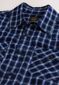 Chemise en flanelle à carreaux bleus avec col, boutons pression et deux poches poitrine, en tissu doux et texturé.