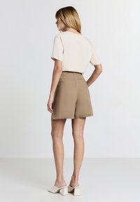 Haut beige à manches courtes associé à un short taille haute beige. Le short a une texture lisse et des poches arrière. Mules à talons carrés blancs.