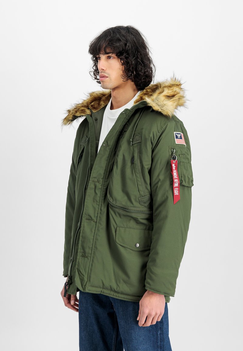 Ung mann iført en olivengrønn parka med faux pels hette, militærstil lapper og et rødt "Remove Before Flight"-merke på lommepocket.
