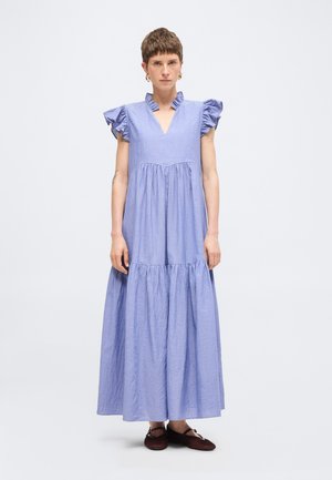 Frau mit kurzen Haaren, die ein hellblaues Maxikleid mit Rüschen an Ärmeln und Kragen trägt, kombiniert mit dunklen flachen Schuhen, vor einfachem Hintergrund stehend.