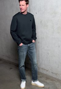 Mann in schwarzem Pullover, blauen Jeans und weißen Sneakern steht mit den Händen in den Taschen vor einer grauen Betonwand.