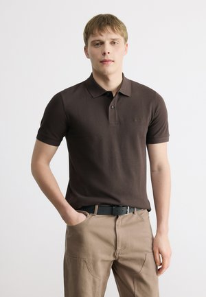 Uomo in piedi con una mano in tasca, indossando una polo marrone scuro e pantaloni beige con una cintura nera su sfondo semplice.