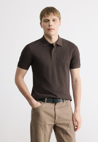 Homme debout, une main dans la poche, portant un polo marron foncé et un pantalon beige avec une ceinture noire, sur un fond uni.