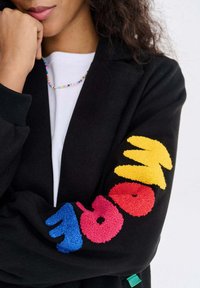 Kobieta ubrana w czarną kurtkę z kolorowymi fakturowanymi literami na rękawie tworzącymi napis "WOW", białą koszulę oraz wielokolorowy naszyjnik z koralików.