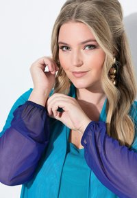 Tyrkysový top s průhlednými fialovými rukávy, staženými na zápěstích. Modelka si dotýká vlasů, viditelné zlaté výrazné náušnice. Jemná, hladká textura.