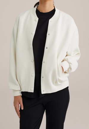 Person iført en åben hvid bomberjakke med knapper over en sort skjorte og sorte bukser, med den ene hånd i jakkelommen.