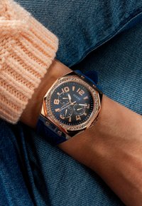 Orologio in oro rosa con cinturino in silicone blu, cassa ottagonale, quadrante nero, grandi numeri e accenti in cristallo. Indossato a un polso.