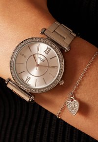 Silberfarbene Uhr mit rundem Zifferblatt, Kristalllünette, römischen Ziffern und metallischem Armband. Begleitet von einem silbernen Herzanhängerarmband.