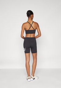 Černá sportovní podprsenka s překříženými ramínky, doplněná černými cyklistickými šortkami s bílým logem. Modelka má na nohou bílé tenisky s texturovanými detaily.