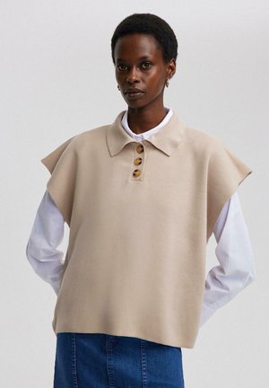 Beige pullover met korte capmouwen, kraag, vier grote knopen, gedragen over een witte blouse, gecombineerd met een spijkerrok.