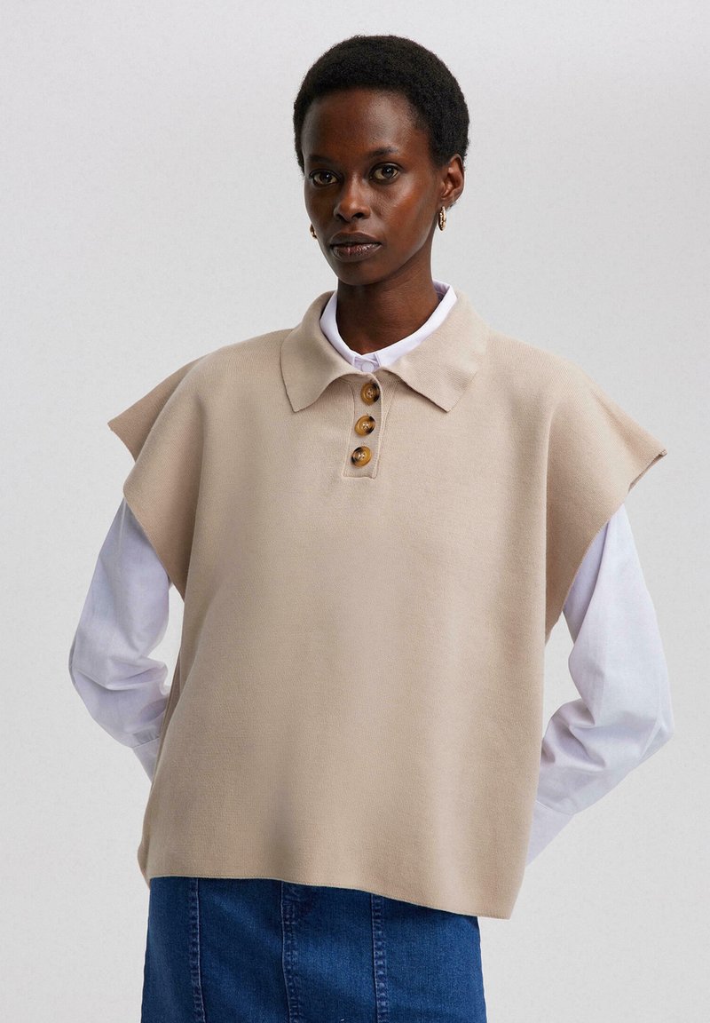 Beige pullover met korte capmouwen, kraag, vier grote knopen, gedragen over een witte blouse, gecombineerd met een spijkerrok.