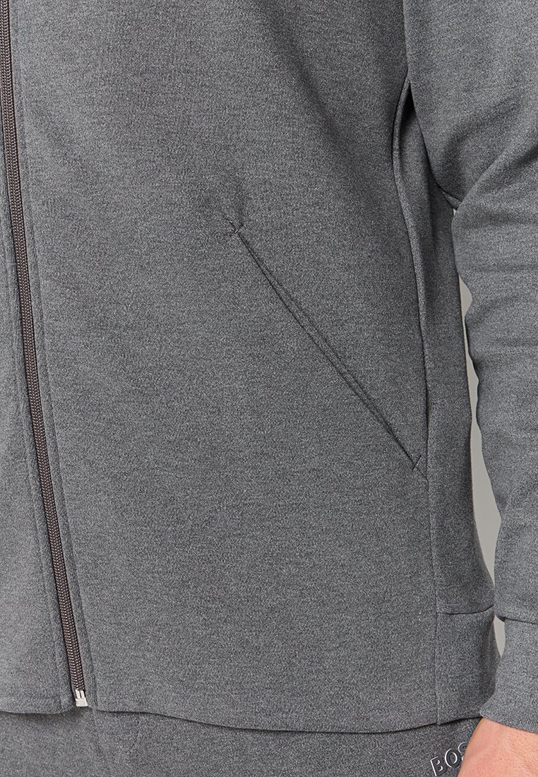 Grijze ritsjack met een gladde textuur, voorzien van een zijzak en gedetailleerde stiksels langs de rits. Slank model.