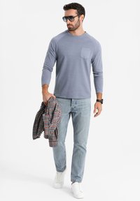 Vaaleansininen pitkähihaisuus paita, jossa on rintatasku, yhdistettynä vaaleisiin farkkuihin. Malli pitää kädessään puna-mustaraidallista paitaa ja käyttää valkoisia tennareita.