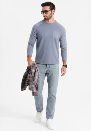 Helles blaues Langarmhemd mit einer Brusttasche, kombiniert mit hellen Jeans. Das Model hält ein rot-schwarzes Karoshirt und trägt weiße Sneakers.