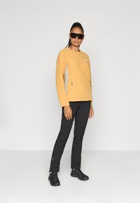 Pulover din fleece galben muștar, cu mâneci lungi și buzunare laterale, asortat cu pantaloni negri slim-fit și încălțăminte sport negre. Ochelari de soare închiși la culoare purtați.