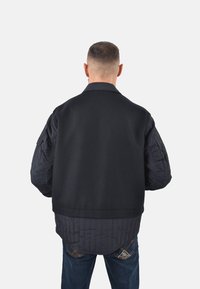 Giacca bomber nera con maniche a righe navy in stile trapuntato e colletto liscio, con una vestibilità comoda e orlo a costine. Indossata sopra jeans in denim.