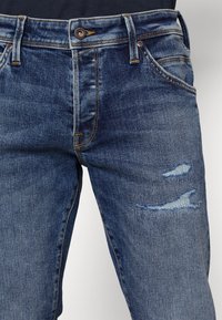 Jeans de mezclilla en azul oscuro con un lavado desgastado, que presentan detalles desgastados en el muslo y costuras amarillas en contraste.