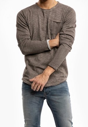 Uomo con camicia marrone a maniche lunghe con taschino sul petto e jeans blu, braccia incrociate in vita su sfondo bianco.