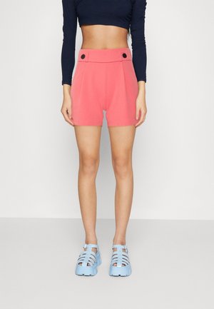 Femme portant un short corail taille haute avec des boutons noirs, un top bleu marine court à manches longues, et des sandales épaisses bleu clair, debout sur un sol gris.