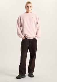Lyserød sweatshirt med rund halsudskæring og lille sort logo, parret med brune bukser med brede ben og grønne sneakers.