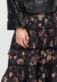 Χέρι ακουμπισμένο σε σκούρο φλοράλ πολύστιχο φόρεμα με βολάν, φορώντας μανικέτα μαύρου δερμάτινου μπουφάν με λεπτομέρεια κουμπιού.