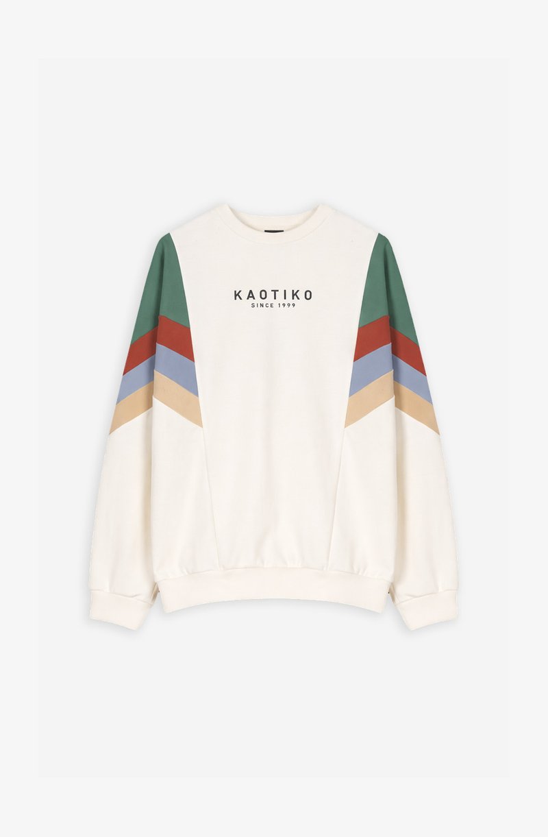 Balta džemperis ar zaļām, sarkanām, zilām un bēšām chevron svītrām uz piedurknēm un tekstu "KAOTIKO SINCE 1999" uz krūtīm.