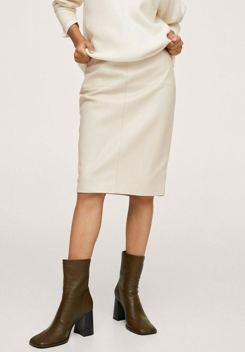 Femme portant une jupe en cuir crème jusqu'aux genoux, un pull ivoire et des bottines à talons en cuir marron, debout les mains ajustant son pull.