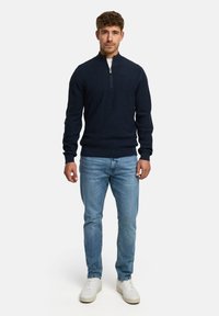 Marineblauer Zip-Knopf-Pullover mit gerippten Bündchen und Saum, kombiniert mit hellblauen Jeans und weißen Sneakers. Der Stoff wirkt weich und strukturiert.