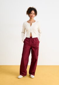 Cremfarbener gerippter Cardigan mit einem Reißverschluss vorne, kombiniert mit lockeren kastanienbraunen Cargo-Hosen mit Taschen und einem kleinen Logo sowie weißen Schuhen.