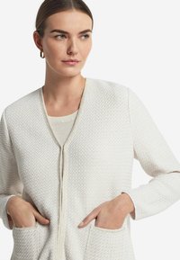 Weißer strukturierter Cardigan mit V-Ausschnitt und Reißverschluss vorne. Verfügt über zwei Fronttaschen und lässige lange Ärmel. Glatte Innenfutter.