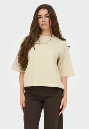 Femme aux cheveux longs et ondulés portant un haut beige surdimensionné à manches courtes avec le texte « DREAM » et un pantalon marron foncé sur un fond uni.