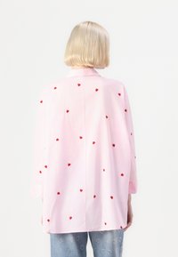 Ροζ πουκάμισο με κόκκινες καρδιές, μακριά μανίκια και κολάρο. Το στρίφωμα έχει ίσια κοπή, σχεδιασμένο για χαλαρή εφαρμογή.