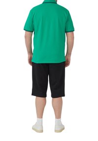 Groene poloshirt met zwarte kraagrand, gecombineerd met zwarte shorts. Model draagt witte sneakers en enkelhoge sokken. Eenvoudig ontwerp.