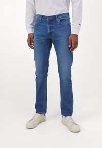 Modré džínové kalhoty s přímým střihem a viditelným prošíváním kapes. Nošeny s bílou košilí s dlouhými rukávy a krémovými teniskami.