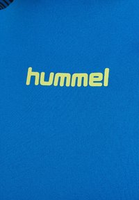Niebieska koszulka sportowa z teksturowanej tkaniny, z wyraźnym żółtym logo "hummel" umieszczonym na środku klatki piersiowej.