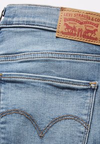 Jasnoniebieskie dżinsy z denimu z brązową skórzaną metką z czerwonym tekstem i logo. Charakteryzujące się wyróżniającym się ściegiem tylnej kieszeni w zakrzywionym wzorze.