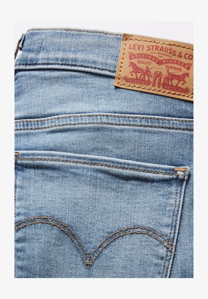 Lichtblauwe denim jeans met een bruine leren label voorzien van rode tekst en een logo. Kenmerkende stiksels op de achterzak in een gebogen patroon.