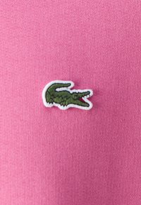 Tessuto rosa con un logo di coccodrillo ricamato in verde, bordato di bianco, posizionato nell'angolo in basso a sinistra.