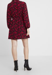 Robe noire à motif floral rouge, taille cintrée, manches longues et jupe évasée. Une paire de bottines noires à talons hauts avec détails de lacets.